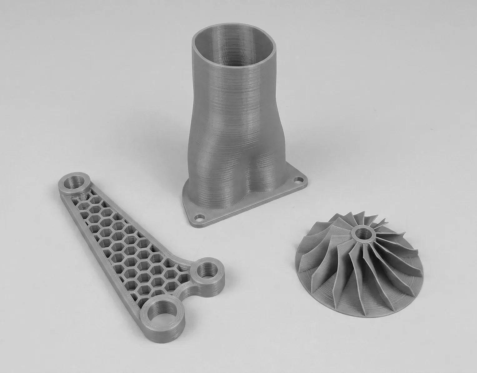 Three parts printed in FDM, in grey: a curved duct with a drilled flange on which the layer lines can be seen, a flat honeycomb-lightened bracket and an impeller with curved blades.